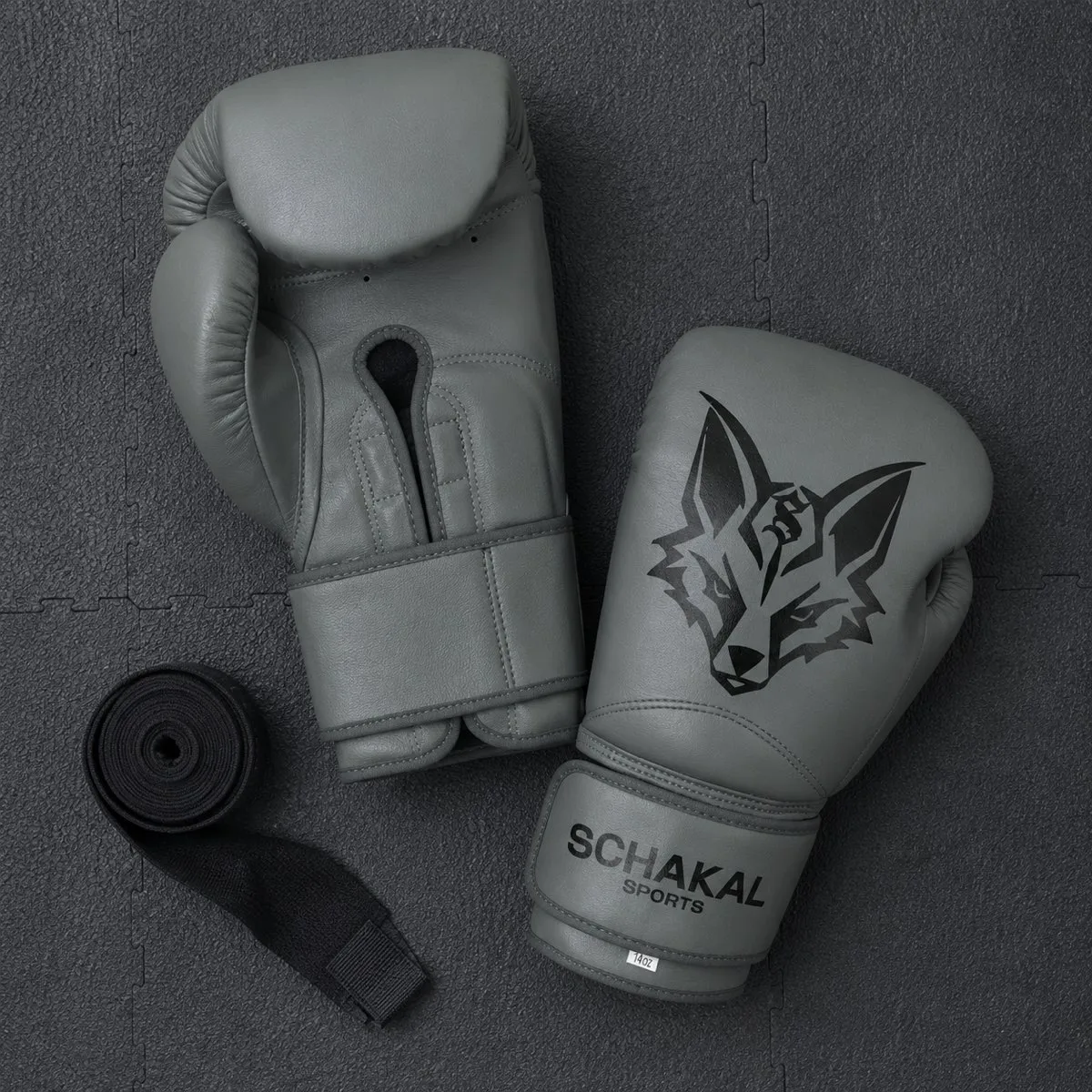 Graue Schakal-Boxhandschuhe von oben auf dunkler Mattenfläche, daneben eine aufgerollte Bandage